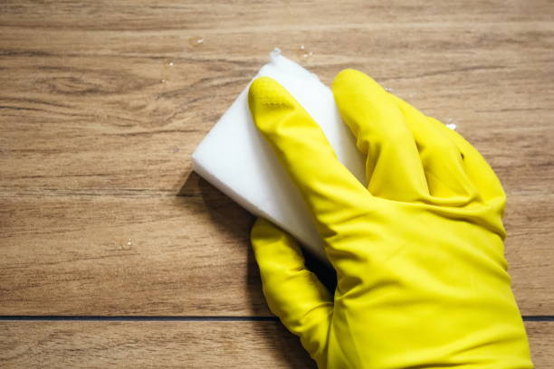 una mano en un guante de goma amarillo se limpia la baldosa húmeda imitando una superficie de madera con una esponja de melamina blanca. limpieza en un baño moderno. enfoque selectivo - chores wood wet indoors fotografías e imágenes de stock