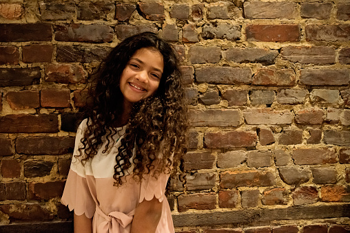 Portrait of beautiful preteen girl. She is from African-American ethnicity, looking at the camera with a smile in front of a brick wall. Horizontal indoors waist up shot with copy space.