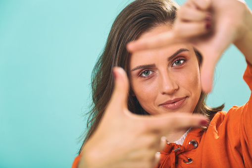Content woman holding finger to create a frame for a photo. Finger framing the composition. Colorful portraits. Blue - cyan background.