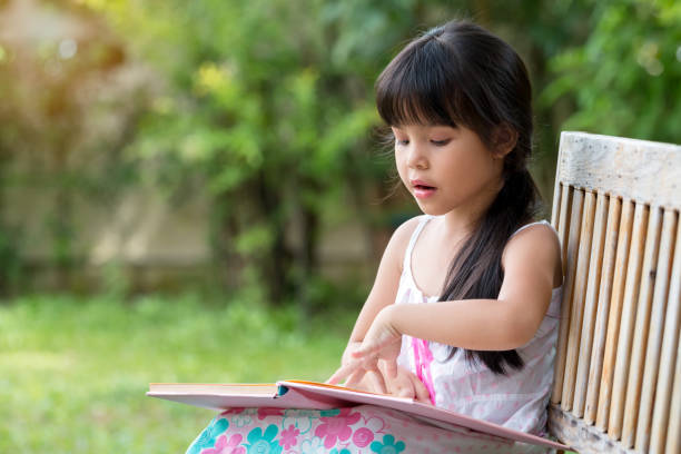 istock Cute Asian girl looking down and reading book while sitting in public Park 1187399611
