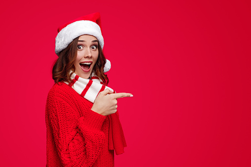 Shocked bright brunette in bright red knitted sweater and Santa hat and scarf pointing with finger away looking amazed at camera on red background