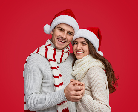 Amorous man and cheerful woman bonding in festive red Christmas hat and smiling with wide smile holding by hands looking at camera
