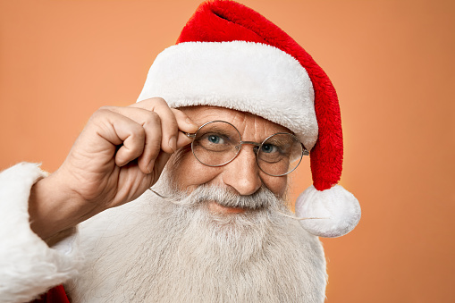 Portrait of senior kind Santa Claus with grey beard and wrinkles on his face looking right to the camera and holding one hand on glasses with orange background. Happy christmas time