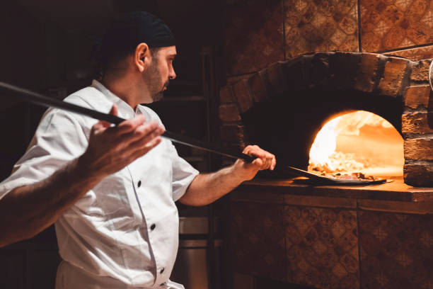 Pizza maker sliding the pizza of a peel in a wood fired oven stock photo