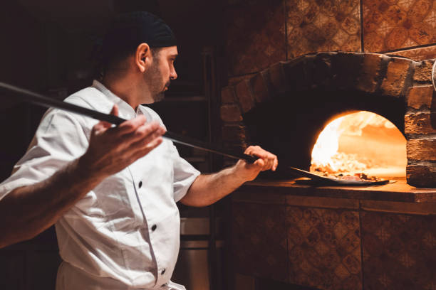 pizza maker sliding the pizza of a peel in a wood fired oven - pizzeria imagens e fotografias de stock