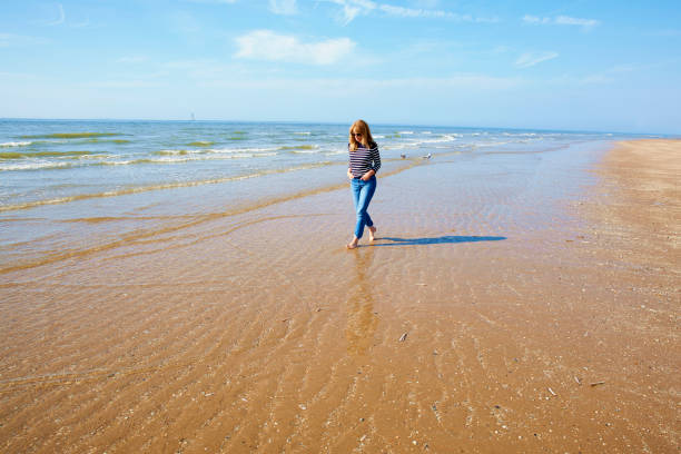 Full length shot of barefoot woman wearing casual clothes and sunglasses while walking on the seaside.