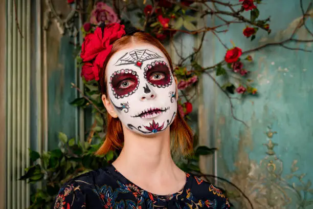 Portrait of calm female celebrates Day of Death, has dark circles near eyes, painted smile, wears traditional mexican attire. People preparing for popular carnival, applied makeup in shape of skull