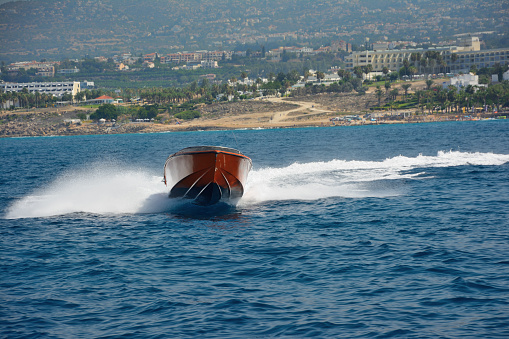 A white yacht sailing on a blue ocean. The yacht is seen moving towards the right side of the image, leaving a trail of white foam behind it. It features a red lifebuoy on the side. In the background, another yacht can be observed, also in motion, heading towards the left side of the image. The ocean appears choppy with small waves, while the sky above is clear and blue. The horizon is visible in the background.