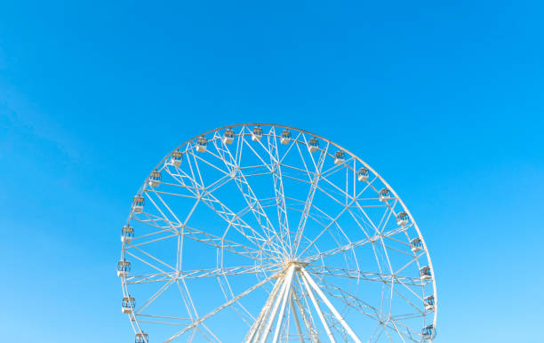 A vibrant ferris wheel with multicolored gondolas stands tall against a bright blue sky, likely in an amusement park setting. The image captures a sense of joy, leisure, and childhood nostalgia. A visible portion of the Sydney Harbour Bridge in the background suggests the photo was taken at Luna Park in Sydney, Australia. Perfect for themes related to fun, travel, holidays, entertainment, and outdoor activities.
