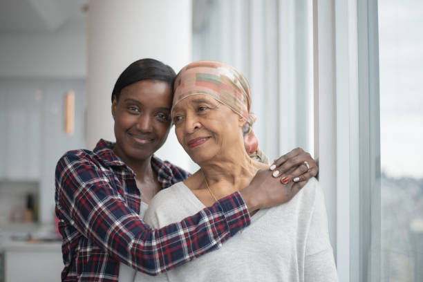 Daughter Hugging Her Mother with Cancer stock photo A young woman of African American decent, stands behind her mother who has cancer, with her arms wrapped around her shoulders hugging her.  Their heads are resting on one another and the senior Mother is wearing a head scarf. scarf photos stock pictures, royalty-free photos & images