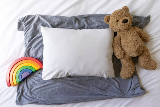 istock Blank white rectangle cushion laying on a grey scarf with a white background with child's teddy and rainbow toy - nursery pillow mockup 1187498037
