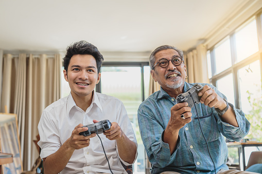 Asian senior father and his adult son playing video games together in living room at home