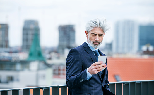 A content mature businessman standing on a terrace, using smartphone.