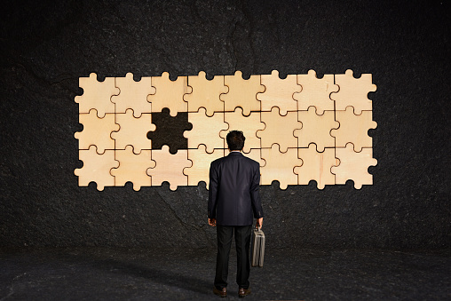 A businessman carrying a briefcase looks at a puzzle hanging on a wall with one of it's pieces is missing