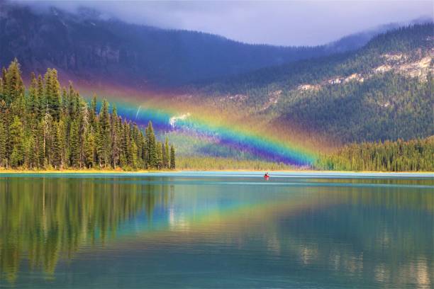 Rainbow ist above on red Kayak on Emerald Lake in Yoho Nationalpark, Western Canada. In the Background the Canadian Rocky Mountains covered with some Clouds. Suns coming out and is shining on the Trees beneath the lake.