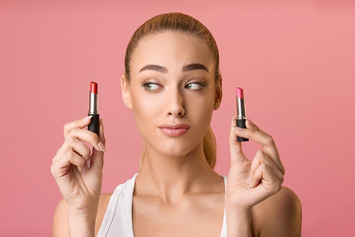 Difficult choice. Girl choosing between two lipstick colors, looking at red lipstick over pink background