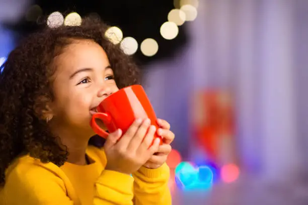 Photo of Cute little african girl with cup of hot cocoa near Christmas tree