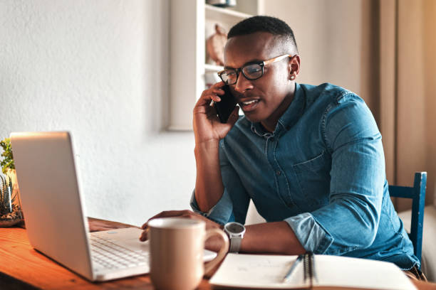 Hello, can I help you? Cropped shot of a handsome young businessman sitting alone in his home office and talking on his cellphone black man laptop stock pictures, royalty-free photos & images