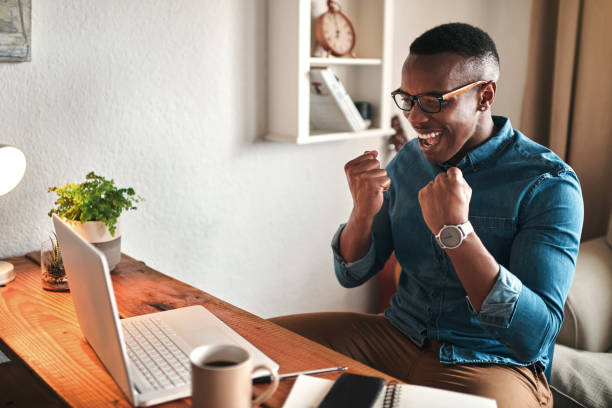 Yes! I did it Cropped shot of a handsome young businessman sitting alone in his home office and feeling excited while using his laptop excitement laptop stock pictures, royalty-free photos & images