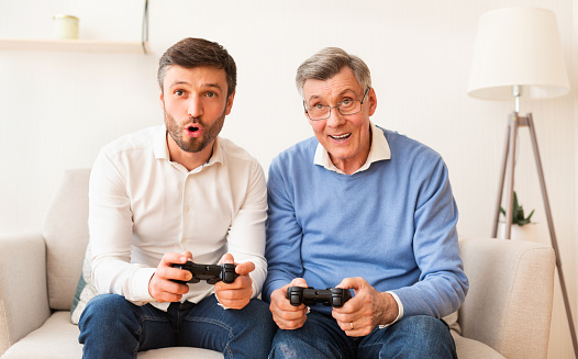 Mature Son And Elderly Father Playing Video Game Having Fun Together Sitting On Sofa Indoor. Different Generations Friendship