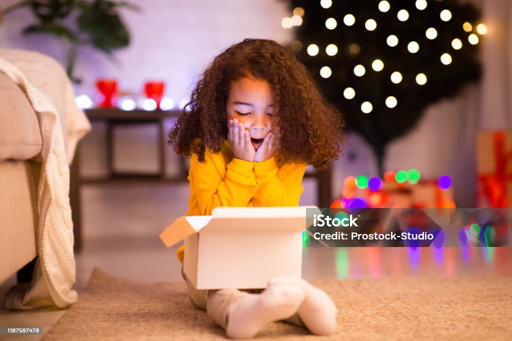 Amazed little afro girl opening Christmas gift near xmas tree Desired present. Amazed little afro girl opening Christmas gift near xmas tree, free space Child Stock Photo