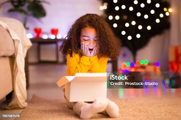 Amazed Little Afro Girl Opening Christmas Gift Near Xmas Tree Stock Photo - Download Image Now