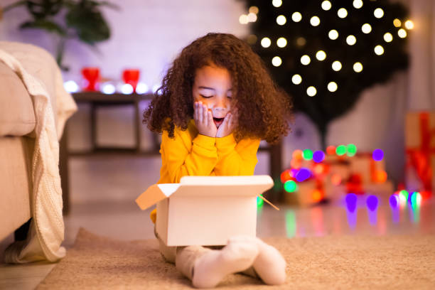 amazed little afro girl opening christmas gift near xmas tree - prenda de natal fotos imagens e fotografias de stock