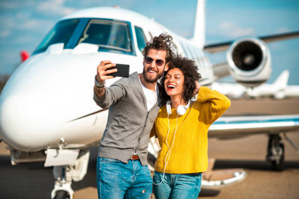 空港のターマックに駐車したプライベートジェット機の前で携帯電話で自分撮りを撮る金持ちの若いカップル - high society audio ストックフォトと画像