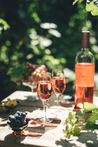 degustazione di vino di rosa, bicchiere di vino di rosa versato dalla bottiglia all'aperto in giardino partito in vigna, uva matura su tavolo di legno, luce del sole, tempo di raccolta, spazio di copia - vino rosato foto e immagini stock