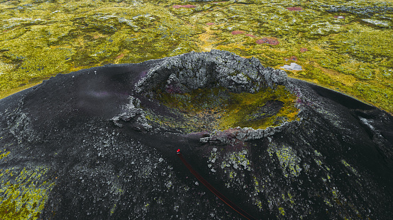 Drone photo of couple in red jackets staying in the crater of a big greeen-black volcano looking at lava fields on Snaefellsnes Peninsula in West Iceland