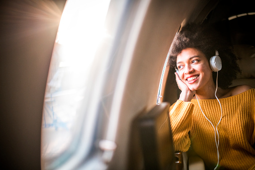 Young chic young woman sitting in a private jet and listening to music through the headphones. She is looking through the window and smiling.