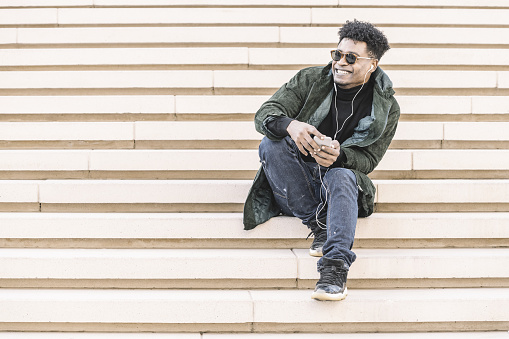 young black man with sunglasses in casual modern clothes sitting on stairs, listen to music of his phone in earphones while smile, technology and lifestyle concept