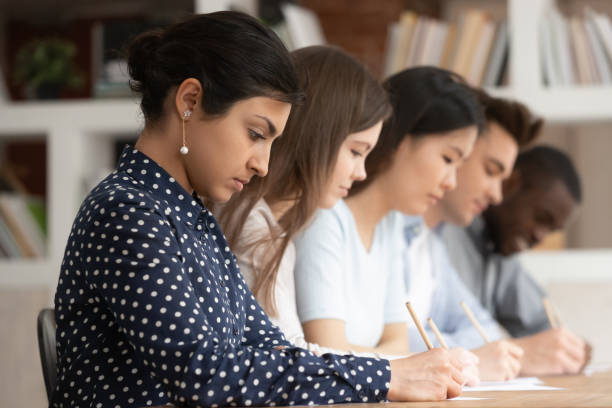 creative writing club images Multiracial students sitting at desk writing on papers doing task Multi-ethnic concentrated students sitting at desk holding pencils writing on papers doing task, diminishing perspective focus on indian girl study process or assessment - knowledge evaluation concept writing workshop stock pictures, royalty-free photos & images
