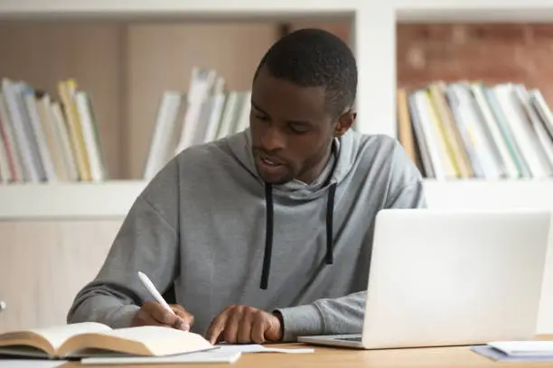 Focused on learning american fellow do schoolwork or preparing for university exams, african guy sitting at classroom desk holding pen writing thinking creating essay or summary work activity concept