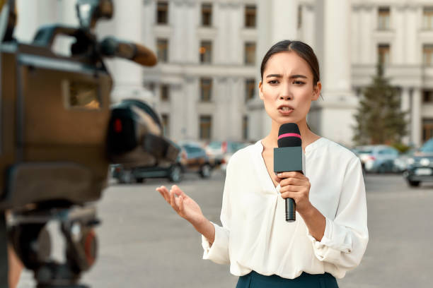 The whole truth and nothing but the truth. TV reporter presenting the news outdoors. Journalism industry, live streaming concept. Cropped portrait of professional female reporter at work. Young woman standing on the street with a microphone in hand and smiling at camera. Horizontal shot. Selective focus on woman newsreader stock pictures, royalty-free photos & images