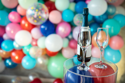Two champagne glasses and a bottle on glass table in room with multi colored balloons