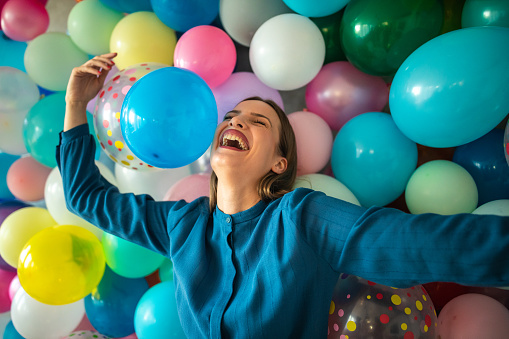 In front of wall with multi colored balloons standing woman in dress and laughing