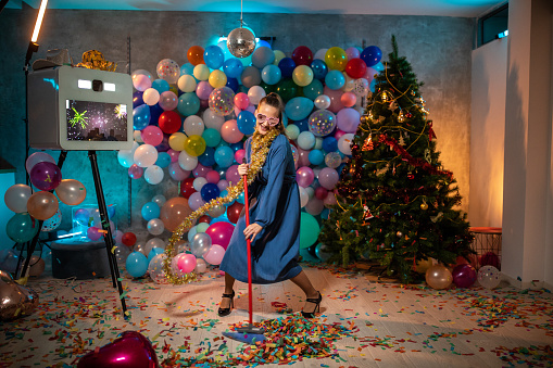 Crazy young woman with broom, in high heels and with gold tinsel decorative tape around neck dancing at messy room