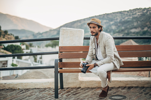 Handsome, bearded man with hat and white, casual suit sitting on bench and holding digital photo camera.