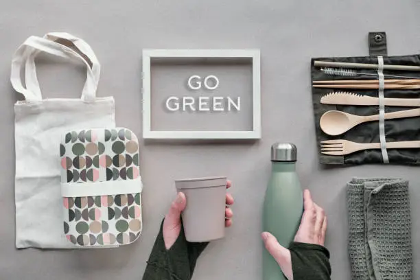 Creative top view, zero waste packed lunch concept. Flat lay, takeaway lunch set - bamboo cutlery, lunch box, cotton bag and hand with coffee-to-go cup on brown paper. Frame with text "Go green".
