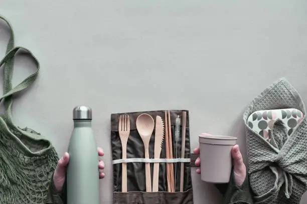 Creative top view, zero waste packed lunch concept, takeaway lunch box set with bamboo cutlery, reusable box, cotton bag and hand with coffee-to-go cup above on craft paper. Sustainable lifestyle.