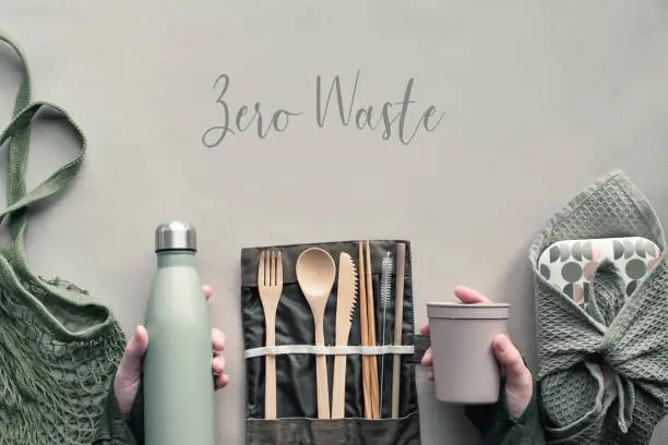 Creative top view, zero waste packed lunch concept. Flat lay, takeaway lunch box set with bamboo cutlery, reusable box, cotton bag and hand with coffee-to-go cup on craft paper. Text "Zero Waste".