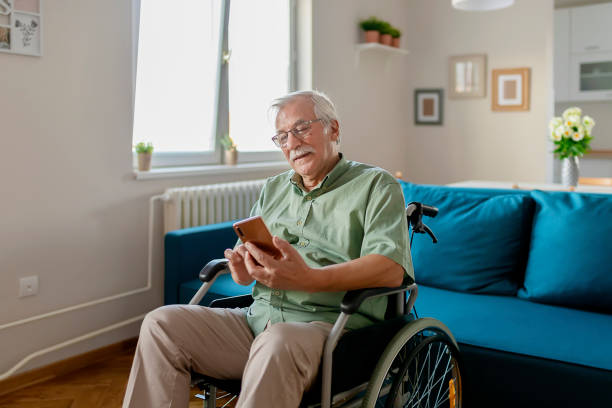 Handicapped Senior Man Sitting in a Wheelchair and Using a Smartphone Handicapped senior person sitting in a wheelchair and using a smart phone at home in the living room web browser photos stock pictures, royalty-free photos & images