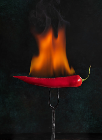 Flaming red pepper on vintage fork fire against dark background. Spicy cuisine concept