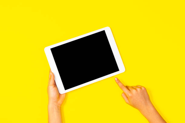 istock Kid hand holding white tablet computer and pointing to blank screen on yellow background 1187687956