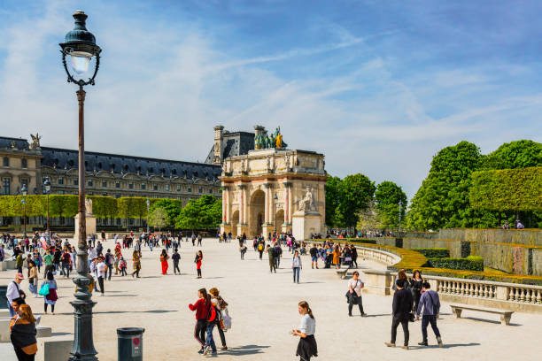 Paris, France - April 18, 2019: Tourists walking on parisian city street in summer. Exterior of old french triumphal arch Arc de Triomphe du Carrousel located in Place du Carrousel. Entrance to Tuileries Garden.