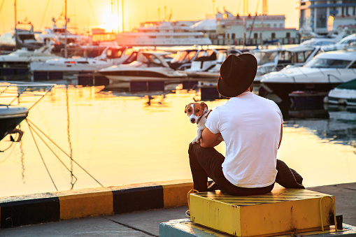 Stylish man and his dog sitting together on pier and enjoying colorful sunset. Man in white t-shirt and black hat. Jack russell terrier puppy on his lap. Luxury yachts docked in sea port.