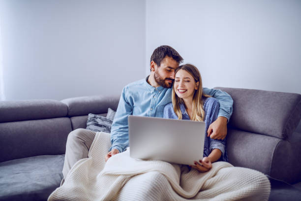 istock Cheerful cute caucasian couple sitting on sofa covered with blanket, snuggling and using laptop for internet surf. Living room interior. 1187690195