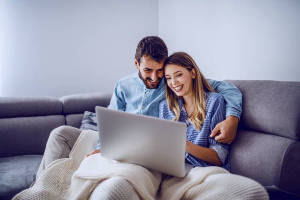istock Cheerful cute caucasian couple sitting on sofa covered with blanket and using laptop for internet surf. Living room interior. 1187690311