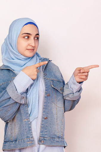 Cute Muslim woman with cheerful face expression points away with her forefinger, showing to copy space on the corner. Look at this. Attractive woman in traditional Arabic clothing points thumb